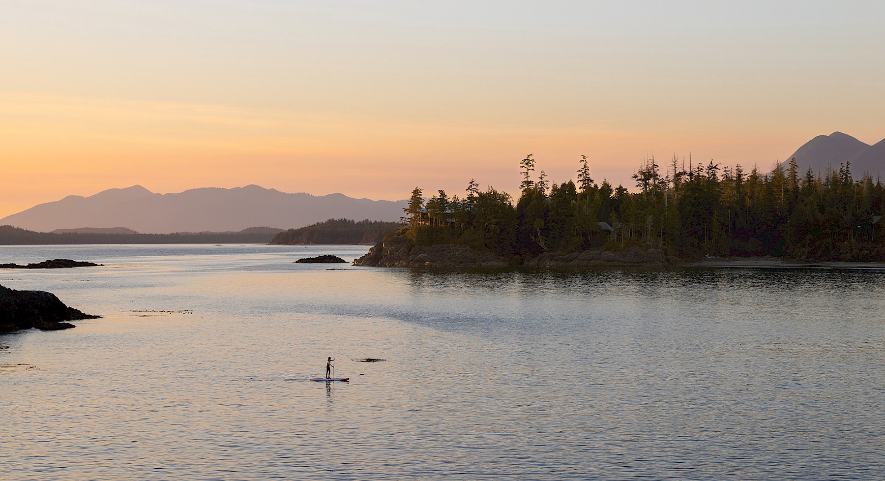A serene sunset view over a calm ocean waters in Tofino, with a person paddleboarding in the center, surrounded by distant mountains and forested islands.