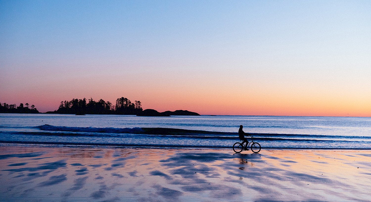 Sunset over a serene beach with a silhouette of a person riding a bicycle along the shoreline, reflecting on the wet sand.