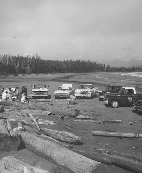 Vintage black and white photo of a group of people gathered around several vehicles and pram on a log-strewn beach with mountains in the background.