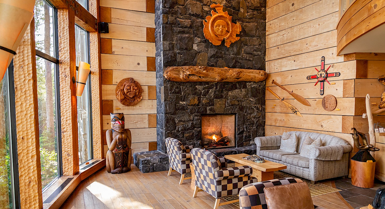 The Beach Lobby at the Wickaninnish Inn with a stone fireplace, carved cedar artwork, and large windows overlooking cedar trees