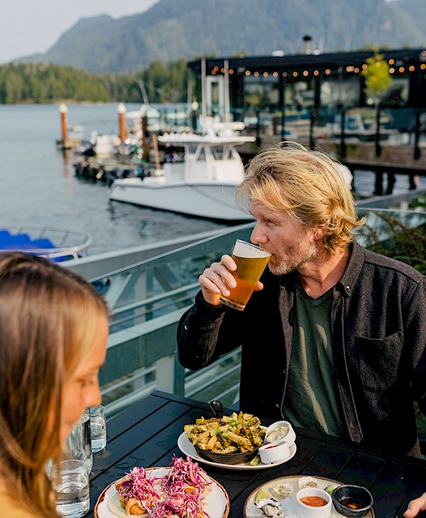 Person enjoying a meal and a drink at an outdoor restaurant by the ocean, with boats and a forested mountain in the background.