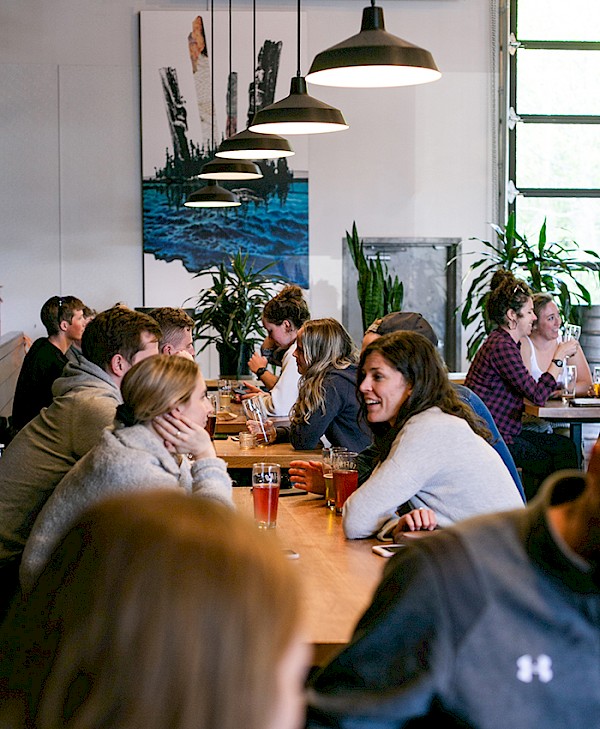 A lively scene inside the Tofino brewery with multiple individuals seated at tables, chatting and enjoying beverages. The decor includes hanging industrial lamps and a large, abstract wall painting.