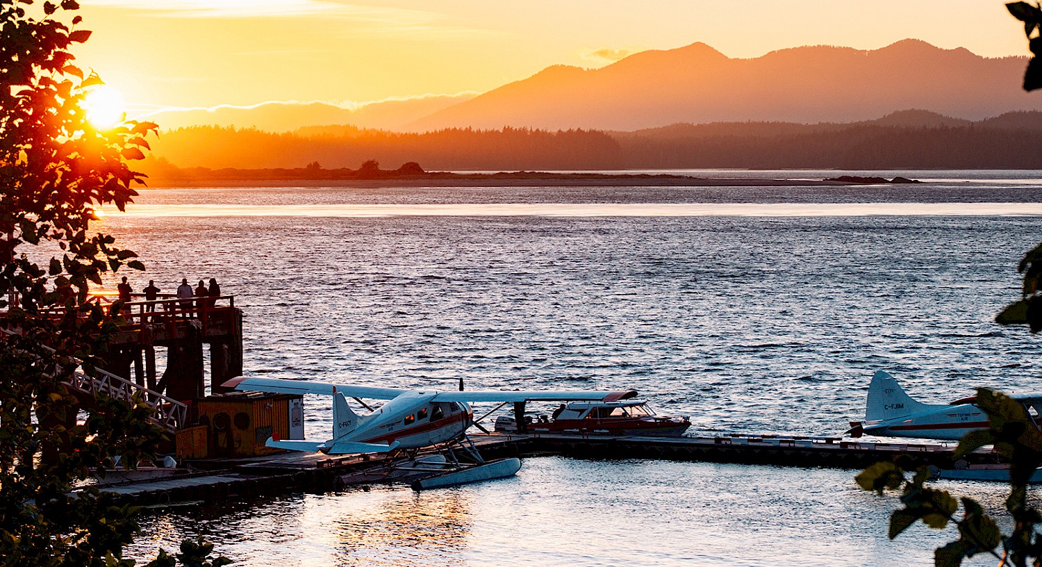 Sunset over a calm inlet with two seaplanes docked, and mountains in the background.