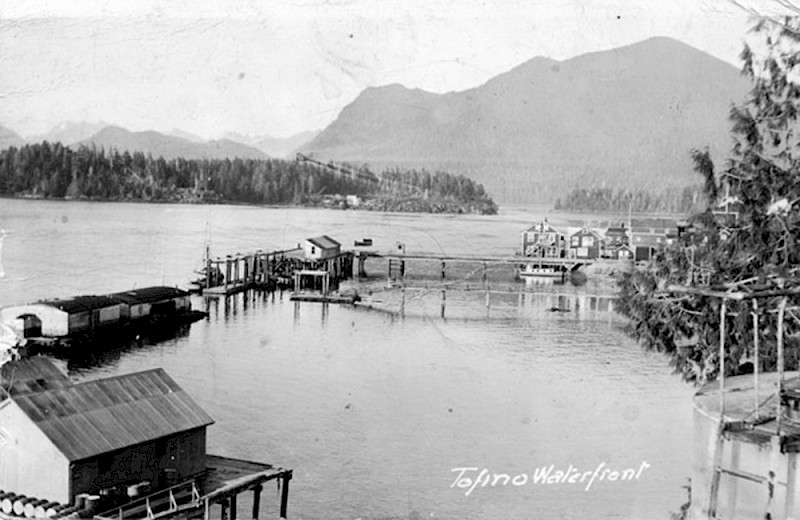 Historic black and white photograph of Tofino Inlet, showcasing a serene ocean flanked by mountains, with floating docks, boats, and buildings
