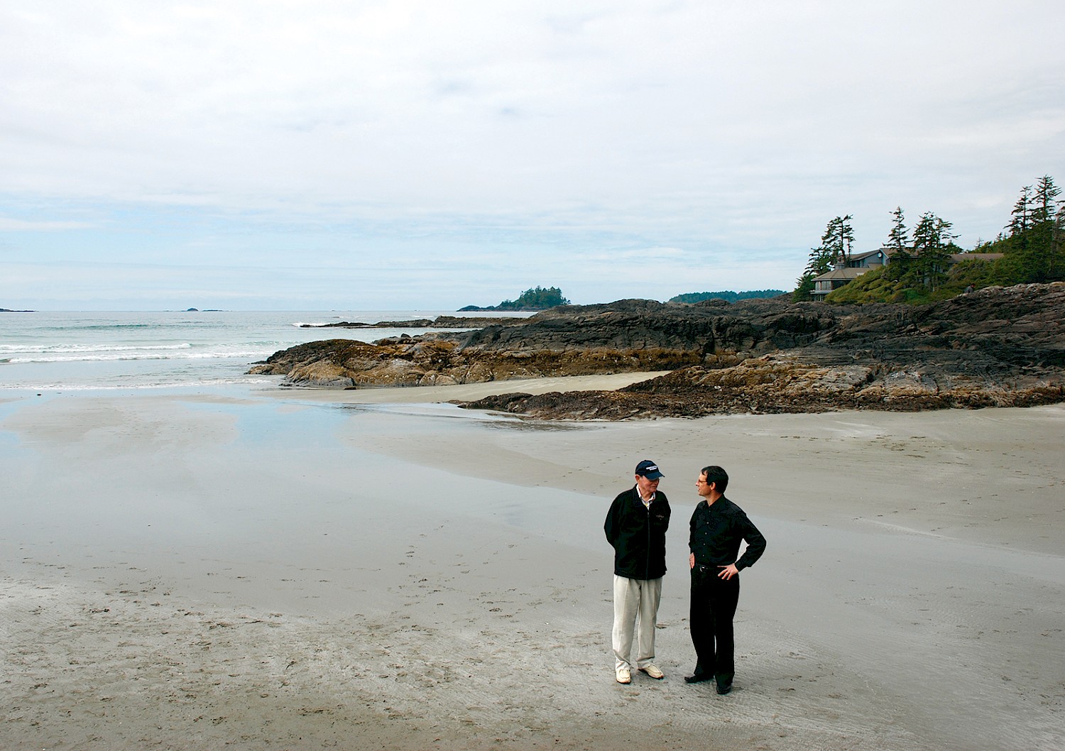 Charles and Howard McDiarmid standing on Chesterman Beach at low tide, conversing, with the ocean, rocky outcrops and Inn in the background.