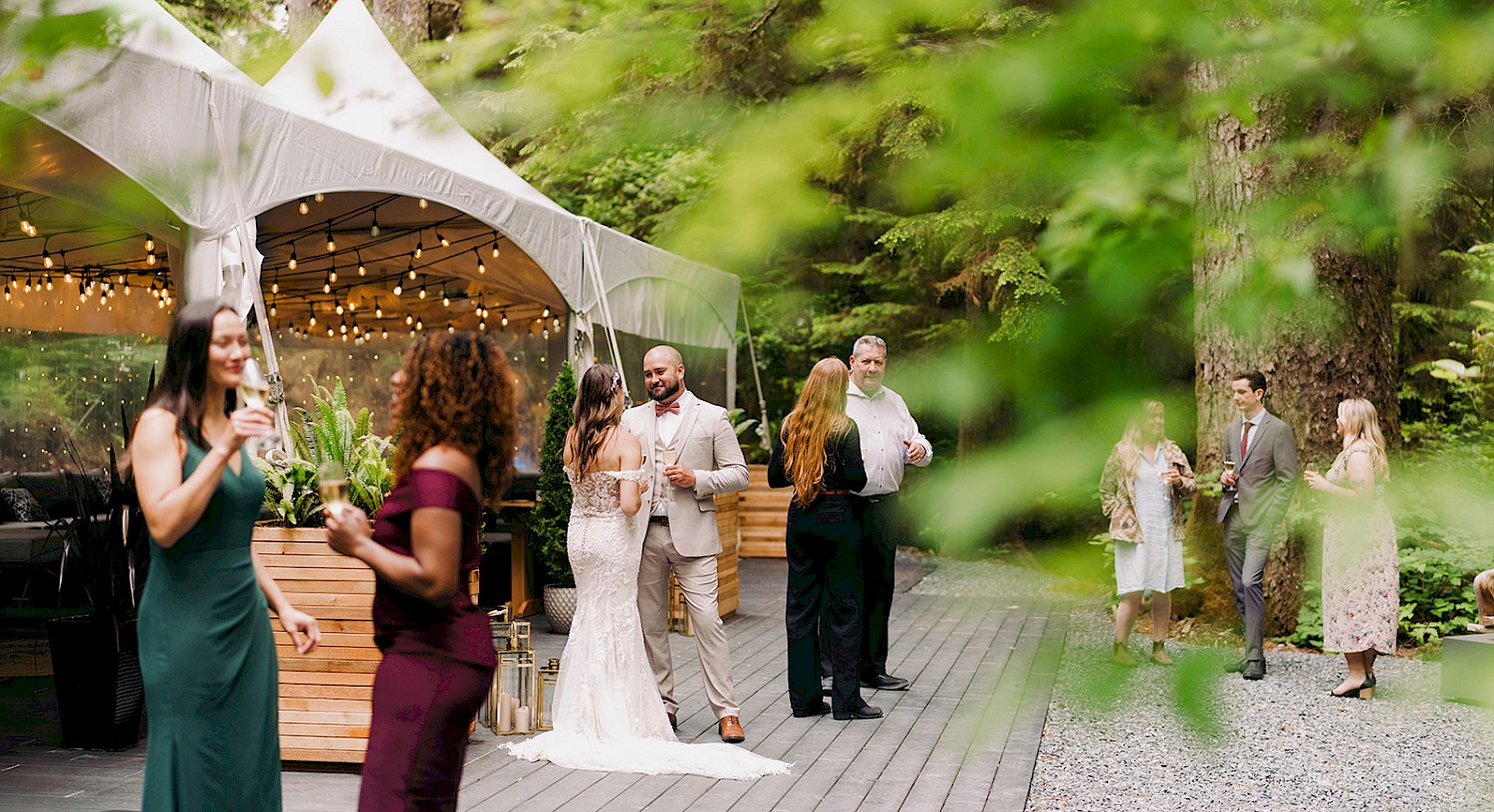 wedding guests gathered in forest event space next to large tent with strung lights