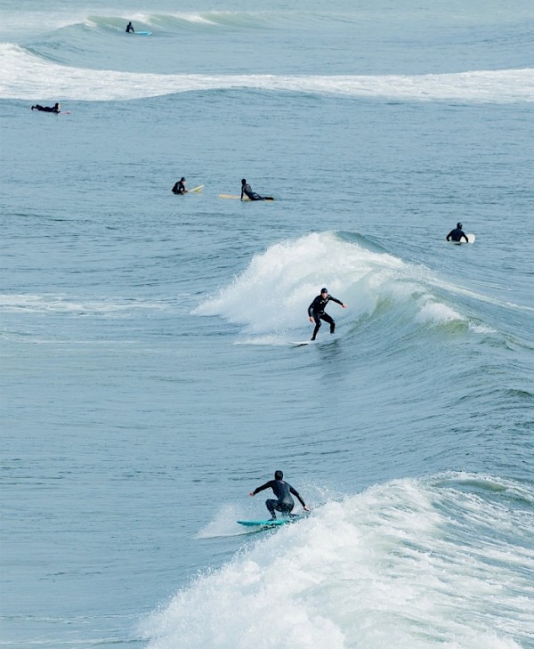 Surfers riding waves in the ocean, with some waiting their turn further out in the water.