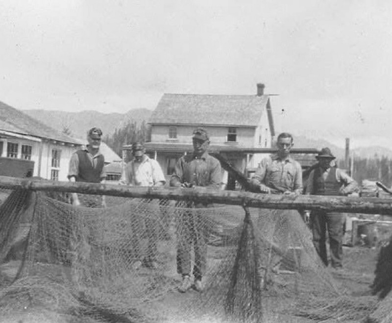 Five individuals standing behind a large fishing net, with rustic buildings and a forested mountain backdrop.