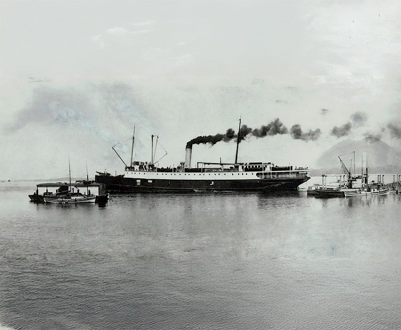 Historic black and white photograph of the SS Maquinna steamship on the water, surrounded by smaller boats, with smoke emitting from its twin funnels.