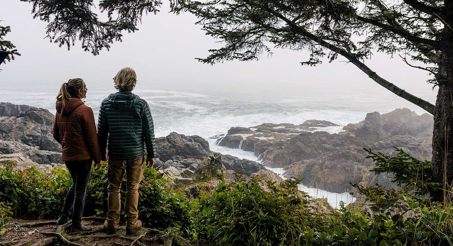 Two people standing on a rocky overlook enjoying the view of a foggy coastline with turbulent ocean waves