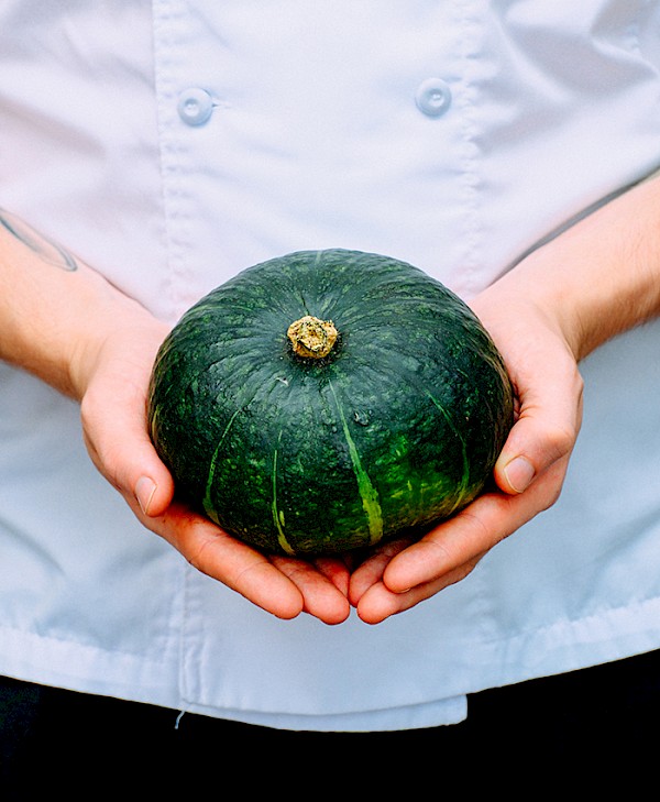 A closeup of hands holding a kabocha squash. The chef is wearing a white chef coat and black pants.