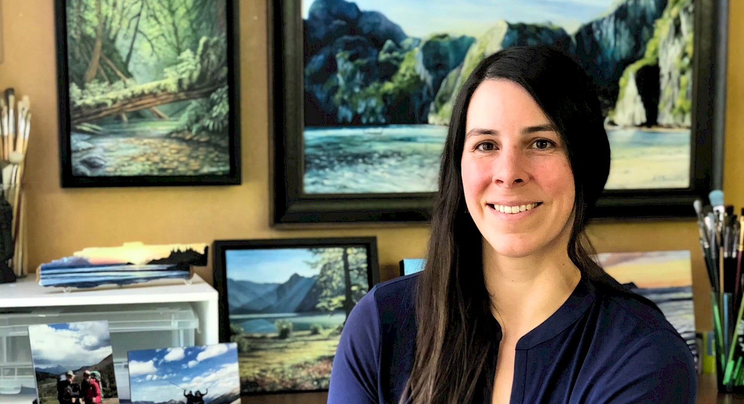 A portrait of Sarah Boileau posing in front of her framed oil paintings hanging on a wall