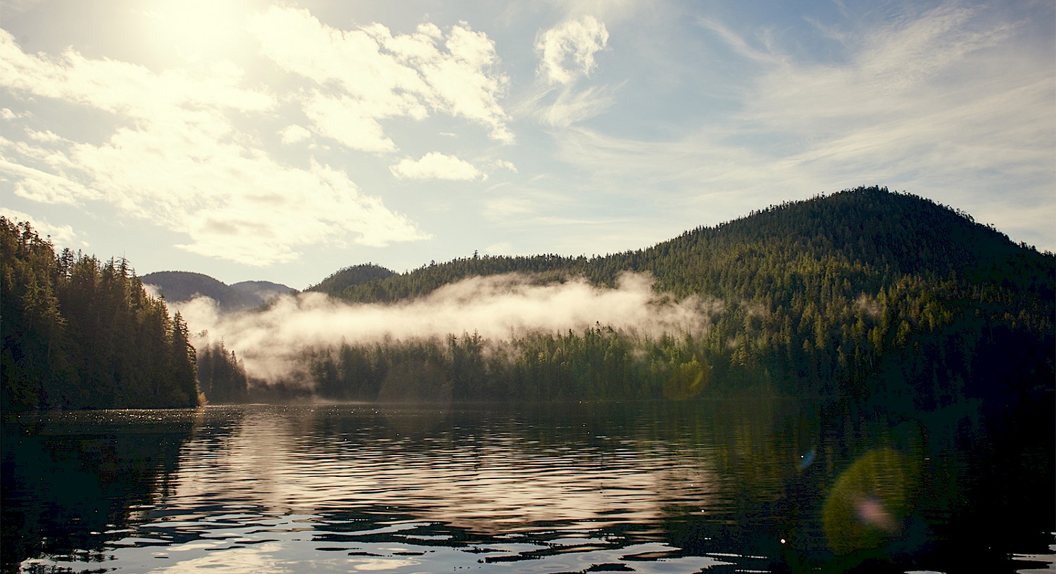 A serene ocean inlet reflecting the surrounding lush, wooded mountains under a partly cloudy sky. A stretch of mist hovers above the water's surface. Sunlight casts a gentle glow over the scene.