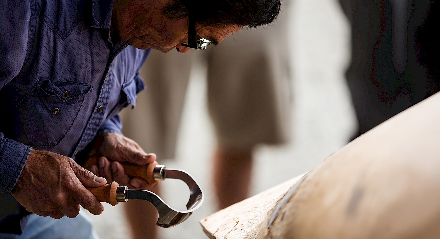A person wearing dark glasses is carving wood with a curved tool, closely focused on their work. The individual is wearing a blue shirt, and there are blurred figures in the background.