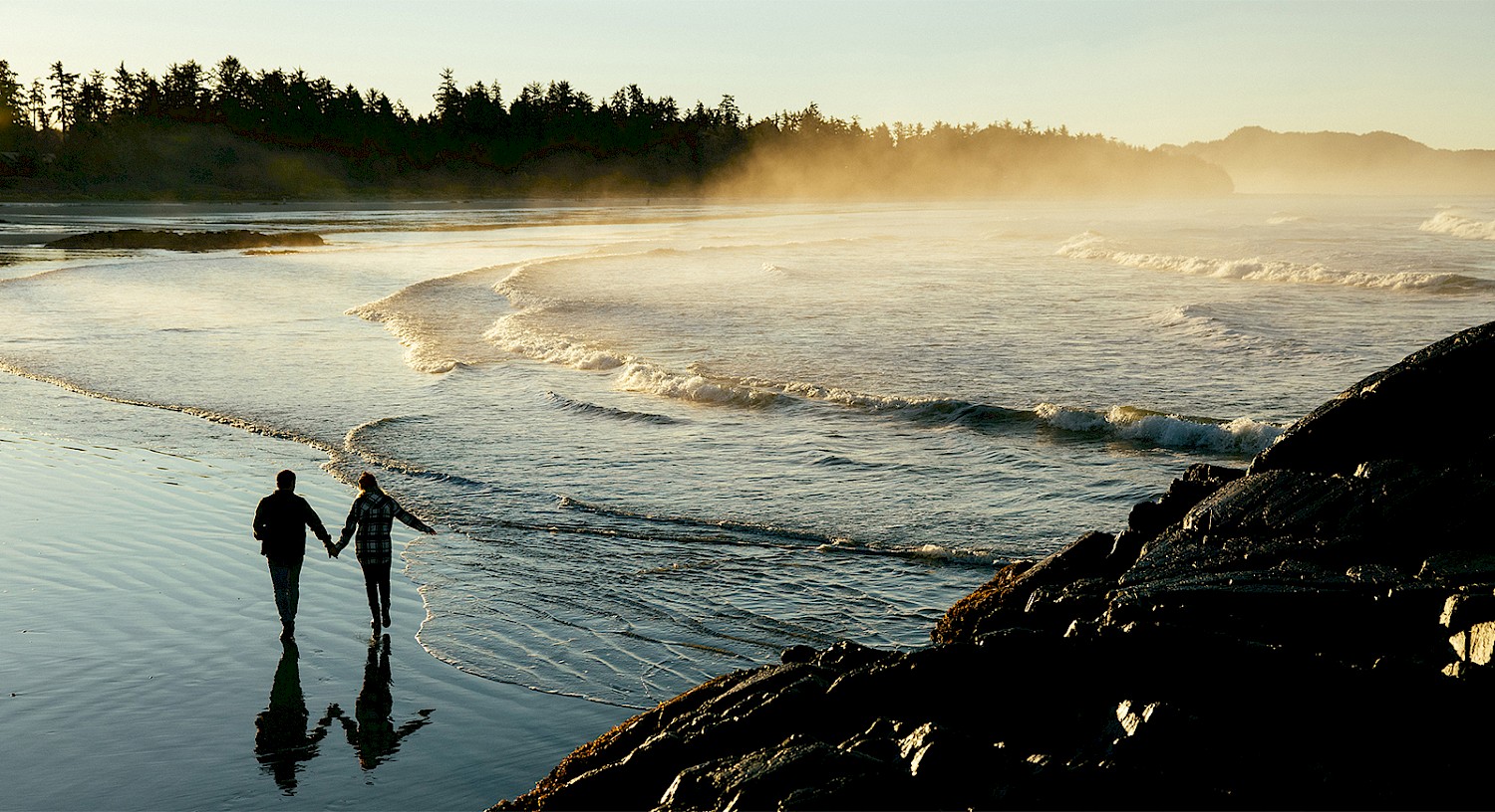 A couple walks hand in hand along the shoreline at a sandy beach during sunrise. Gentle waves wash up against the shore, and a misty haze rises above the distant tree-covered hills. The scene captures a peaceful, romantic moment by the water.