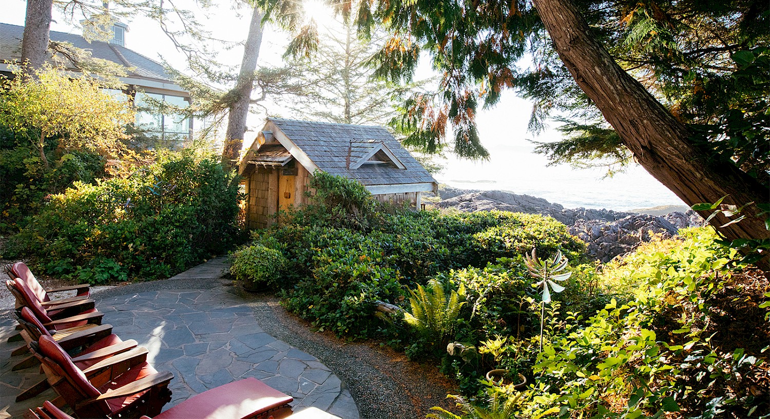 A serene spa patio setting with a small wooden hut surrounded by lush greenery and various trees. Several red lounge chairs are positioned on a stone patio. In the background, there's a view of the rocky shore and the distant ocean under a clear sky.