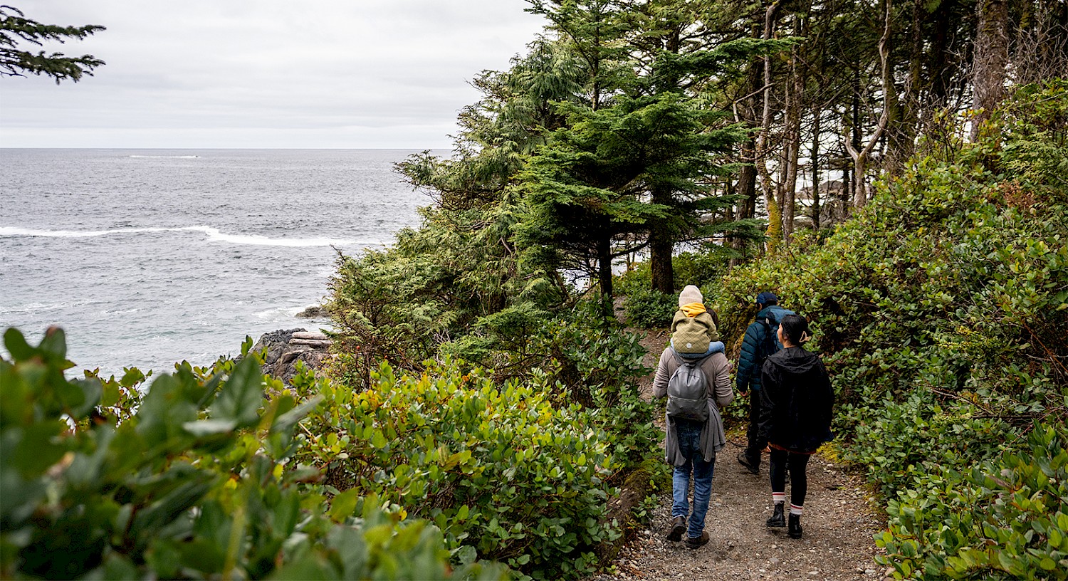 Three adults and a child carried on shoulders hike on a forested coastal trail with the ocean visible through the trees. The path is surrounded by greenery and leads towards a distant shoreline. The sky is overcast.