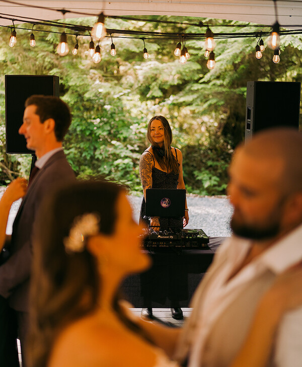 A DJ is playing music outdoors under string lights. Two couples are dancing in the foreground, slightly out of focus. The setting is lush with greenery.