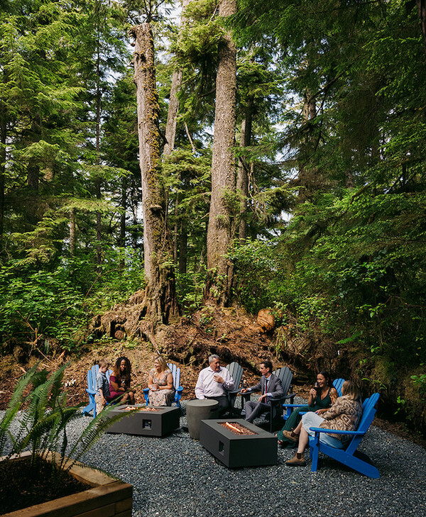 A group of people sit in blue chairs around two modern fire pits in a lush forest setting. Tall trees and dense greenery surround the area, creating a serene and natural environment.