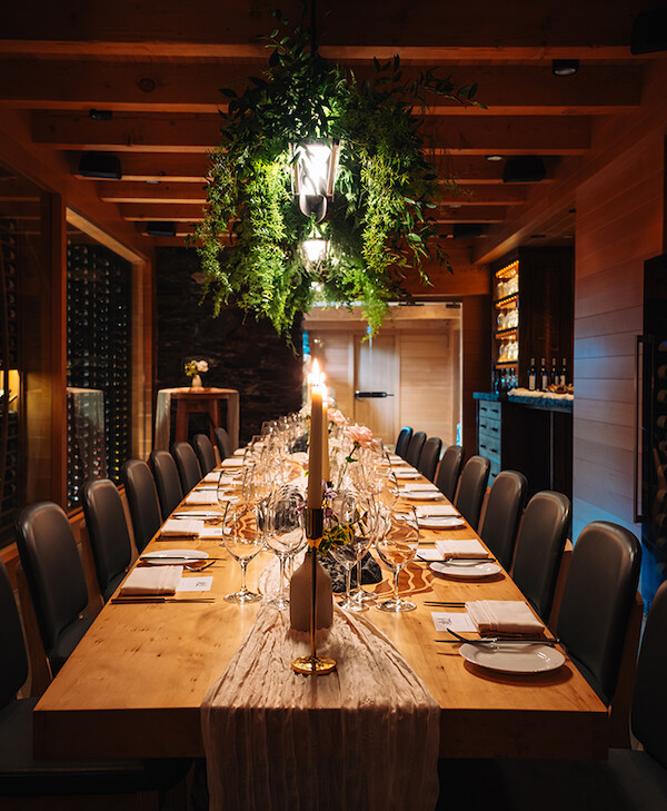 A beautifully set, long dining table is surrounded by chairs in an elegant, warmly lit room. The table is adorned with a white runner, tall candles, and neatly arranged glassware and table settings. A lush green plant arrangement hangs above the table, adding a touch of nature to the sophisticated decor. A wine display and wooden elements in the room contribute to a cozy and refined atmosphere.