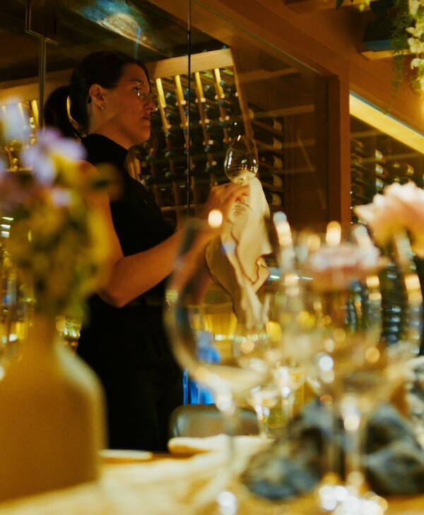 Caroll-Ann polishes a wine glass, rows of wine bottles can be seen behind her and a blurred hint of the table's wedding setup in front