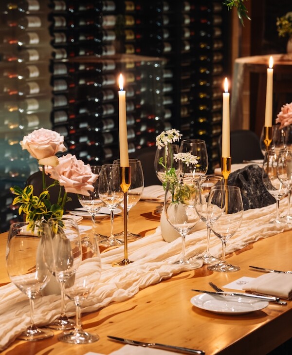 Wine glasses, pink roses, tall candles, silk and table settings beautifully adorn the wooden table with racks of wine bottles in the background