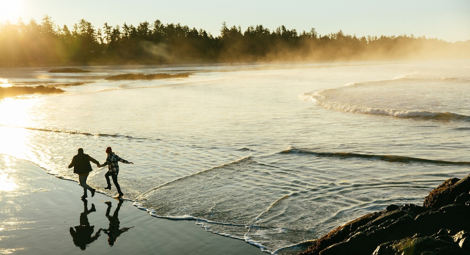 Couple running along Chesterman Beach with mist rolling off the water