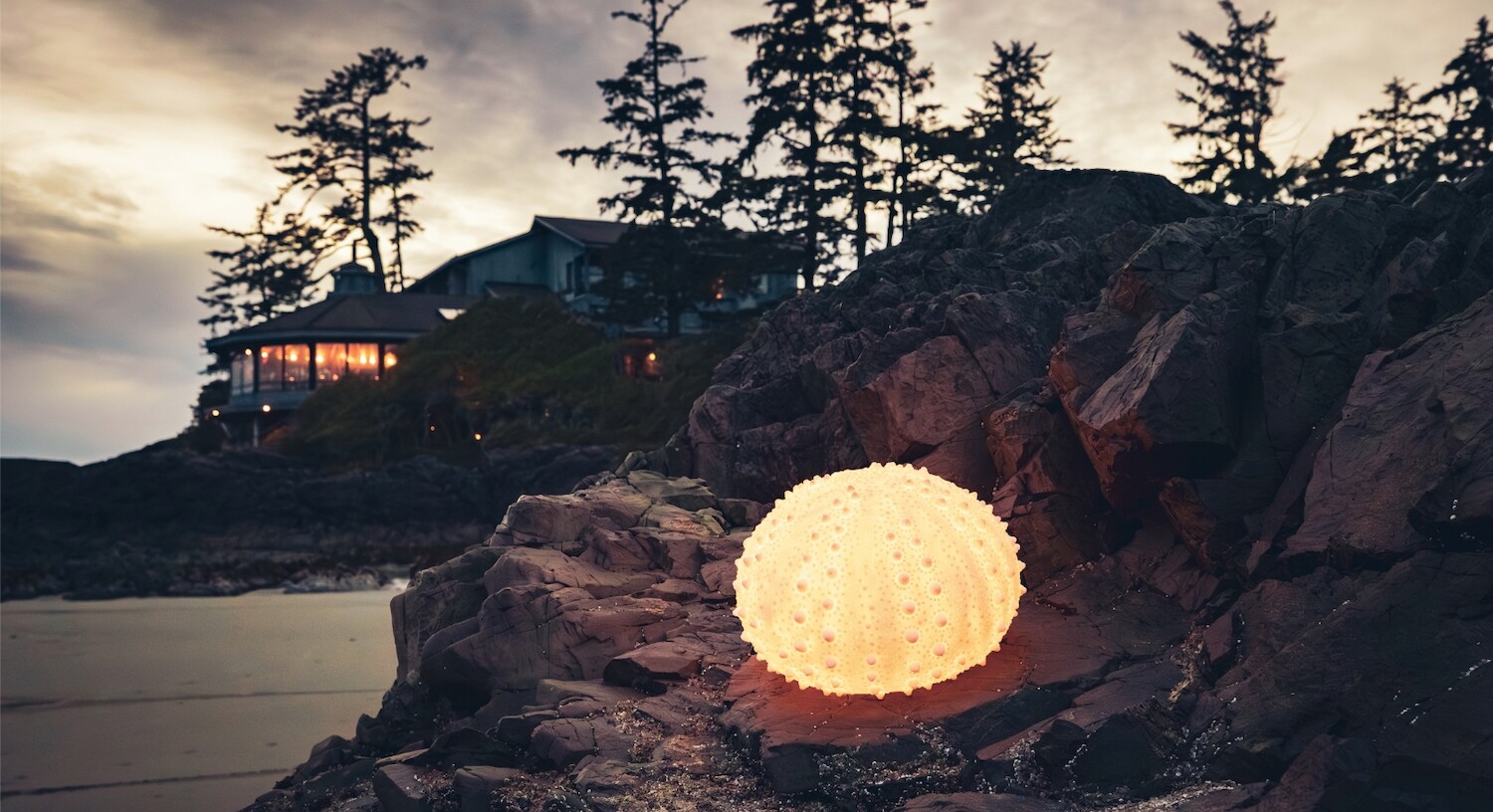 A sea urchin shaped lamp is placed on rocks on Chesterman Beach. The Wickaninnish Inn can be seen in the background.