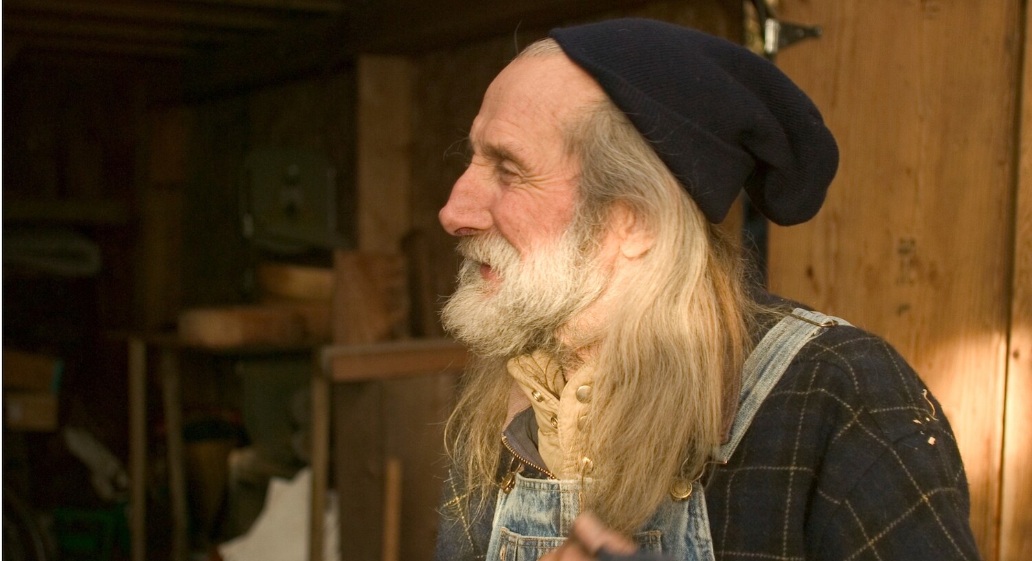 Henry Nolla's side profile, he has long grey hair and a full white beard. He wears a black toque and overalls.