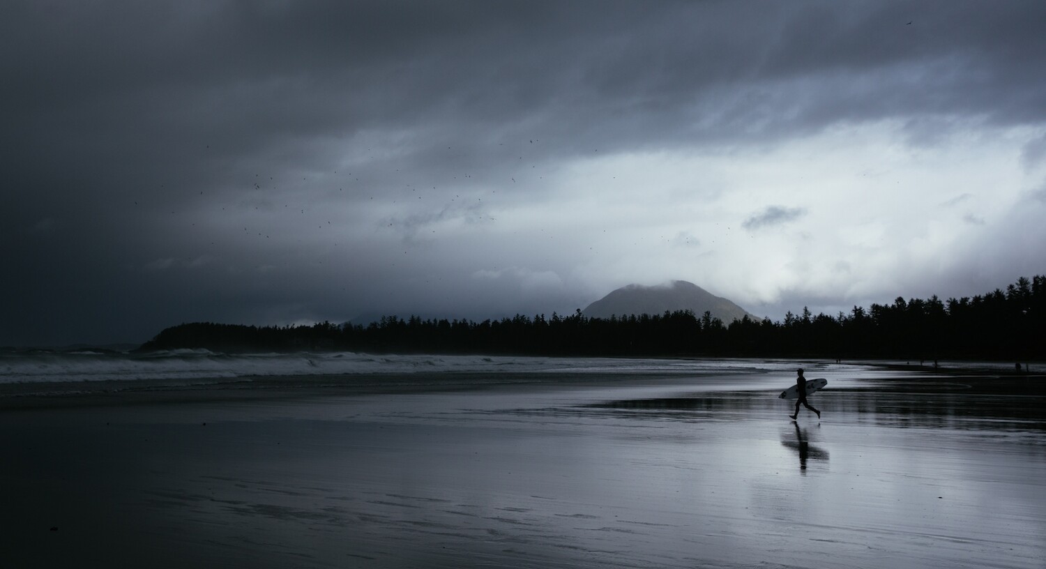 A surfer walks down the glossy sand towards the winter swell on a stark, moody beach in Tofino. In the sky above, a storm gathers - it's eye swirling above Meares Island in the far distance.