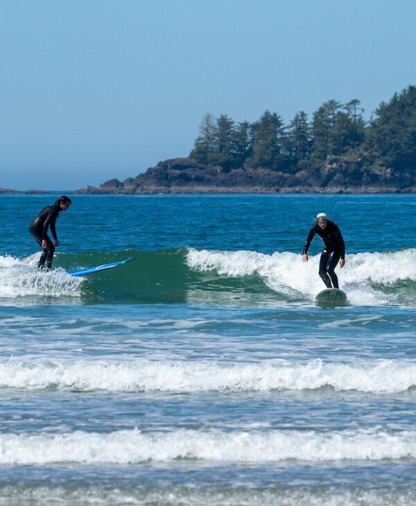 Two surfers ride along a smaller waves of Chesterman Beach, the sky is clear and blue and an island is visible in the distance.